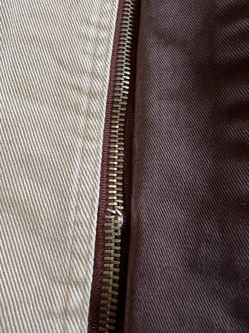 May include: Close-up of a zipper on a garment. The zipper is silver and has a brown fabric backing. The fabric on the left side of the zipper is a light beige color, while the fabric on the right side is a dark brown color.