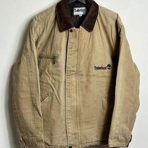 May include: A tan work jacket with a brown corduroy collar and a brown embroidered logo that says "Timberland" on the left chest. The jacket has a zipper closure and two pockets on the front.