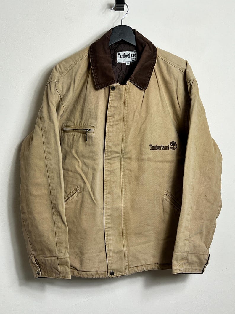 May include: A tan work jacket with a brown corduroy collar and a brown embroidered logo that says "Timberland" on the left chest. The jacket has a zipper closure and two pockets on the front.