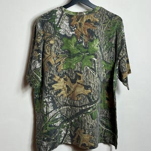 May include: A short-sleeved camouflage t-shirt hangs from a black hanger. The shirt features a pattern of green leaves, brown leaves, and tree bark. The shirt is made of a soft, comfortable material.