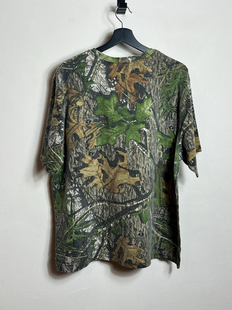 May include: A short-sleeved camouflage t-shirt hangs from a black hanger. The shirt features a pattern of green leaves, brown leaves, and tree bark. The shirt is made of a soft, comfortable material.