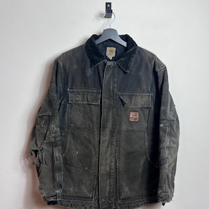 Puede incluir: Una chaqueta de trabajo gris oscuro desgastada con cuello de pana negro cuelga de una percha metálica. La chaqueta tiene dos bolsillos en el pecho, dos bolsillos inferiores y un pequeño parche con el logotipo. La tela muestra signos de desgaste.