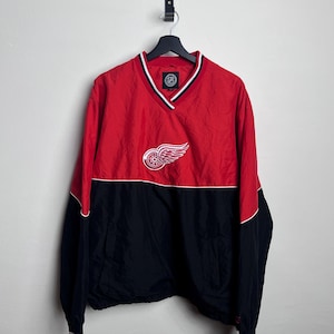 Könnte beinhalten: Rote und schwarze Pulloverjacke mit V-Ausschnitt. Die Jacke hat einen weiß-rot gestreiften Kragen und Manschetten. Ein weißes Logo eines geflügelten Rades befindet sich auf der Brust. Die Jacke hängt an einem schwarzen Kleiderbügel.
