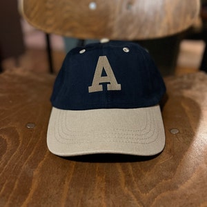 Könnte beinhalten: Eine schwarze und beige Baseballkappe mit einem großen, braunen Filzbuchstaben 'A' auf der Vorderseite.