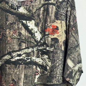 K&ouml;nnte beinhalten: T-Shirt mit Camouflage-Muster und dem Logo "Mossy Oak Break-Up" in Orange. Das Shirt weist eine Mischung aus braunen, gr&uuml;nen und beigen Farben auf, die eine Waldumgebung nachahmen. Das Shirt hat einen Rundhalsausschnitt und kurze &Auml;rmel.