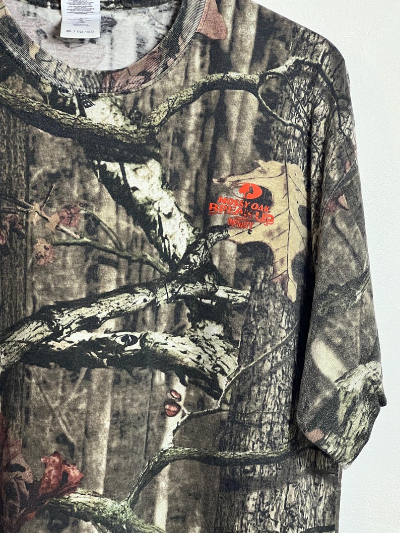 K&ouml;nnte beinhalten: T-Shirt mit Camouflage-Muster und dem Logo "Mossy Oak Break-Up" in Orange. Das Shirt weist eine Mischung aus braunen, gr&uuml;nen und beigen Farben auf, die eine Waldumgebung nachahmen. Das Shirt hat einen Rundhalsausschnitt und kurze &Auml;rmel.