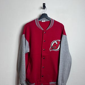 May include: A red and gray varsity jacket with the New Jersey Devils logo on the chest. The jacket has a button-up front, gray sleeves, and striped detailing on the collar, cuffs, and hem. The jacket is hanging on a black hanger.