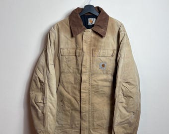 Puede incluir: Chaqueta de trabajo color beige con cuello de pana marrón, dos bolsillos en el pecho y un pequeño parche con el logotipo. La chaqueta está colgada de una percha negra contra una pared blanca. La chaqueta parece desgastada.