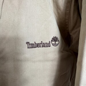 May include: A close-up of a beige jacket with a brown embroidered logo that says "Timberland" with a small tree graphic.