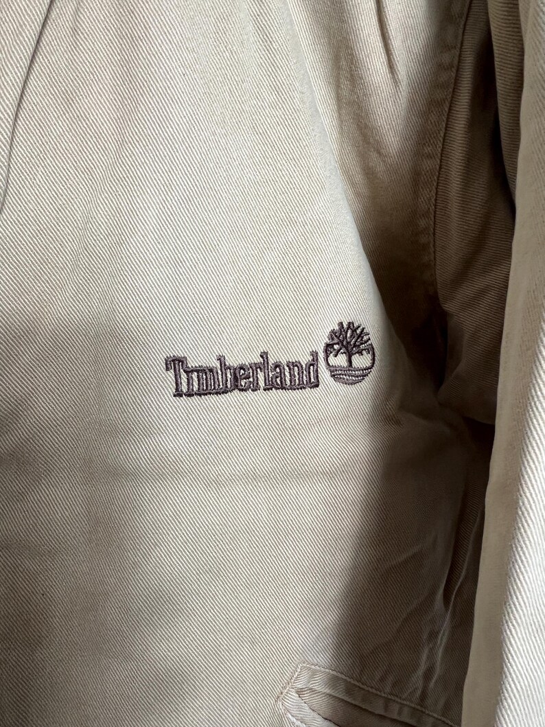 May include: A close-up of a beige jacket with a brown embroidered logo that says "Timberland" with a small tree graphic.