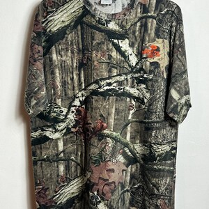 K&ouml;nnte beinhalten: T-Shirt mit Camouflage-Muster und Rundhalsausschnitt. Das Shirt hat ein braunes, gr&uuml;nes und beiges Waldmuster. Das Shirt hat ein kleines orangefarbenes Logo im oberen rechten Brustbereich. Das Shirt h&auml;ngt an einem schwarzen Kleiderb&uuml;gel.