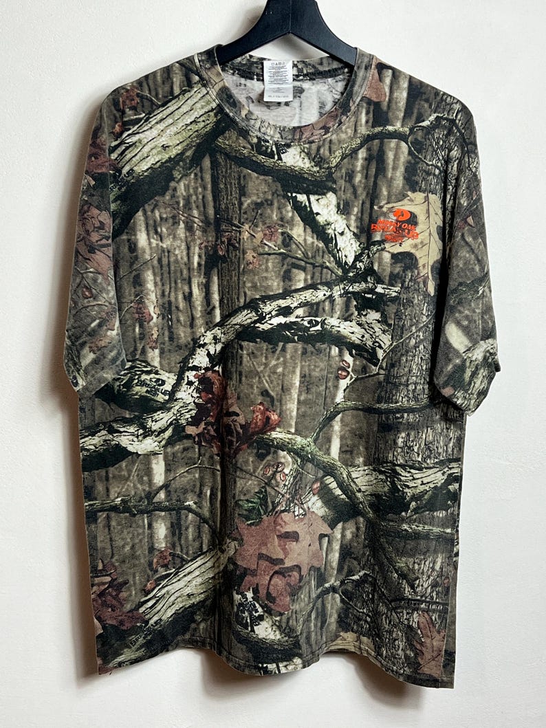 K&ouml;nnte beinhalten: T-Shirt mit Camouflage-Muster und Rundhalsausschnitt. Das Shirt hat ein braunes, gr&uuml;nes und beiges Waldmuster. Das Shirt hat ein kleines orangefarbenes Logo im oberen rechten Brustbereich. Das Shirt h&auml;ngt an einem schwarzen Kleiderb&uuml;gel.