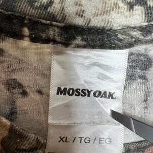 K&ouml;nnte beinhalten: Nahaufnahme eines T-Shirts mit Tarnmuster. Das Etikett des Shirts zeigt den Markennamen "Mossy Oak" in schwarzer Schrift. Das Etikett gibt auch die Gr&ouml;&szlig;e XL/TG/EG an. Der Stoff hat eine Mischung aus Braun-, Gr&uuml;n- und Beiget&ouml;nen.