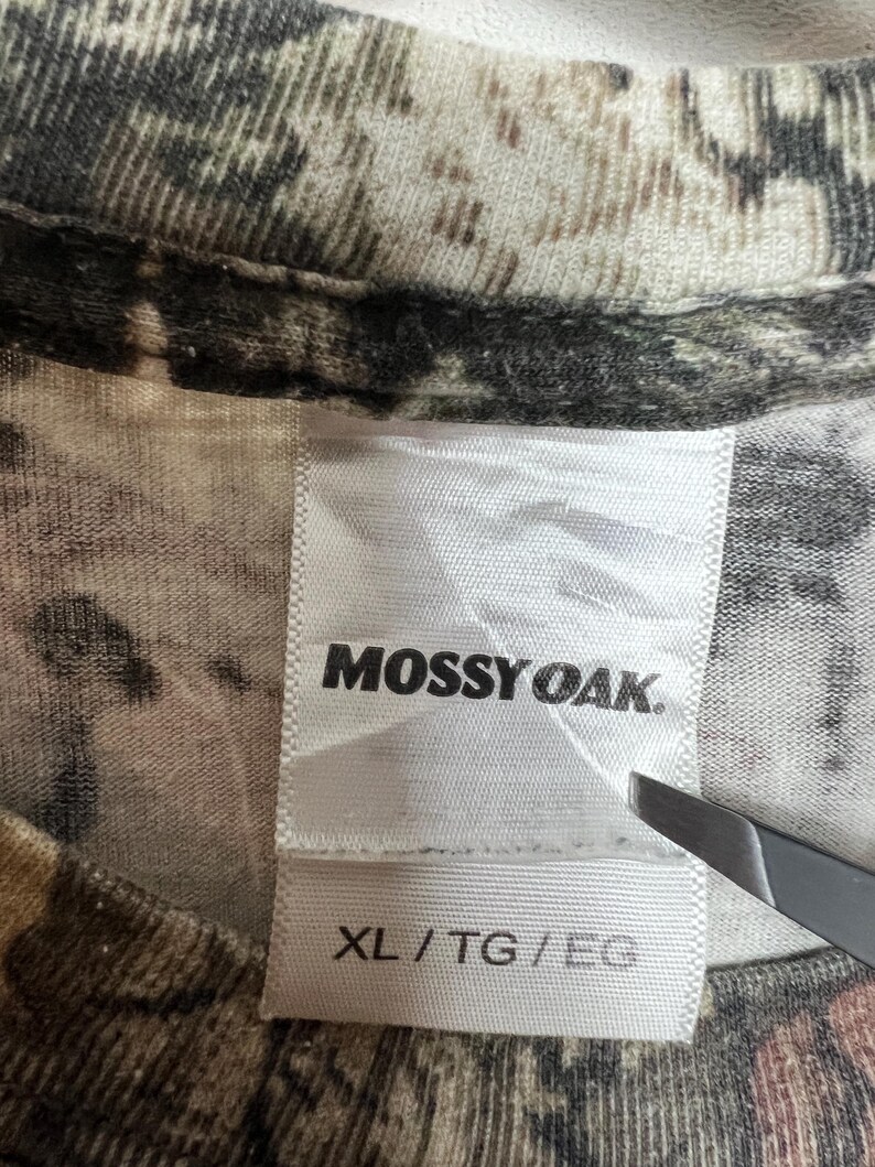 K&ouml;nnte beinhalten: Nahaufnahme eines T-Shirts mit Tarnmuster. Das Etikett des Shirts zeigt den Markennamen "Mossy Oak" in schwarzer Schrift. Das Etikett gibt auch die Gr&ouml;&szlig;e XL/TG/EG an. Der Stoff hat eine Mischung aus Braun-, Gr&uuml;n- und Beiget&ouml;nen.