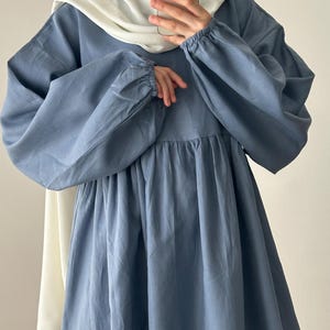May include: A dusty blue long-sleeved dress featuring a gathered waistline and elasticized cuffs, paired with a solid white headscarf. The dress has a loose, flowing silhouette with a round neckline and a soft, matte fabric texture throughout.