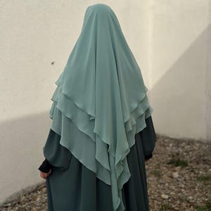 Puede incluir: Una abaya larga y fluida de color verde azulado con un hiyab a juego, de color verde azulado claro y en capas. La abaya tiene mangas largas y un corte holgado y fluido.