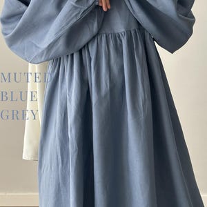 May include: A long-sleeved dress in a muted blue-grey color featuring a gathered waistline and voluminous sleeves with elasticated cuffs. The text MUTED BLUE GREY is printed on the left side of the garment, which also shows a white fabric panel.