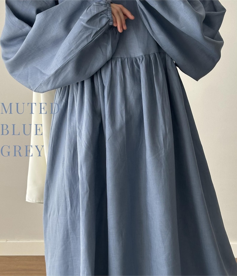 May include: A long-sleeved dress in a muted blue-grey color featuring a gathered waistline and voluminous sleeves with elasticated cuffs. The text MUTED BLUE GREY is printed on the left side of the garment, which also shows a white fabric panel.