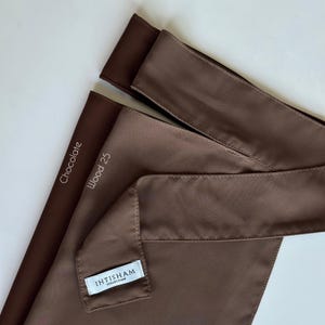 May include: Two brown fabric headbands, one labeled "Chocolate" and the other "Wood 25". The headband labeled "Wood 25" has a small white tag that reads "IHTISHAM COLLECTION".
