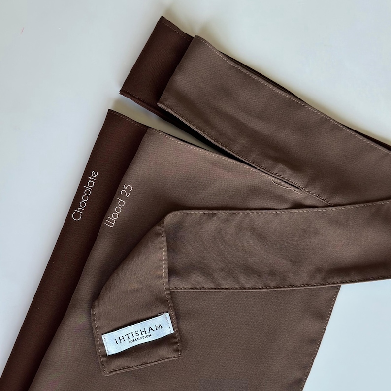 May include: Two brown fabric headbands, one labeled "Chocolate" and the other "Wood 25". The headband labeled "Wood 25" has a small white tag that reads "IHTISHAM COLLECTION".