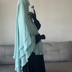Könnte beinhalten: Ein helltürkisfarbener Hijab mit gerüschten Schichten wird über einem schwarzen Kleid getragen. Die Person hält ein weißes Smartphone und trägt schwarze Handschuhe. Der Hintergrund ist eine helle Wand und ein graues Sofa.