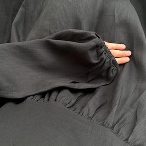 May include: A long-sleeved black dress featuring a gathered waistline and elasticized cuffs on the sleeves. The fabric has a visible woven texture, and the garment is constructed with a full, flowing skirt that drapes downward from the fitted bodice.