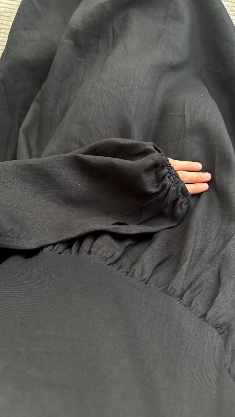 May include: A long-sleeved black dress featuring a gathered waistline and elasticized cuffs on the sleeves. The fabric has a visible woven texture, and the garment is constructed with a full, flowing skirt that drapes downward from the fitted bodice.