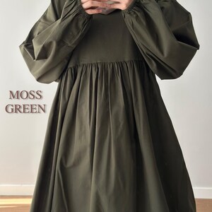 May include: A moss green dress featuring long, voluminous sleeves with elasticated cuffs and a gathered, flowing skirt. The text MOSS GREEN is visible on the left side. The garment has a round neckline and a loose, relaxed fit throughout the bodice.