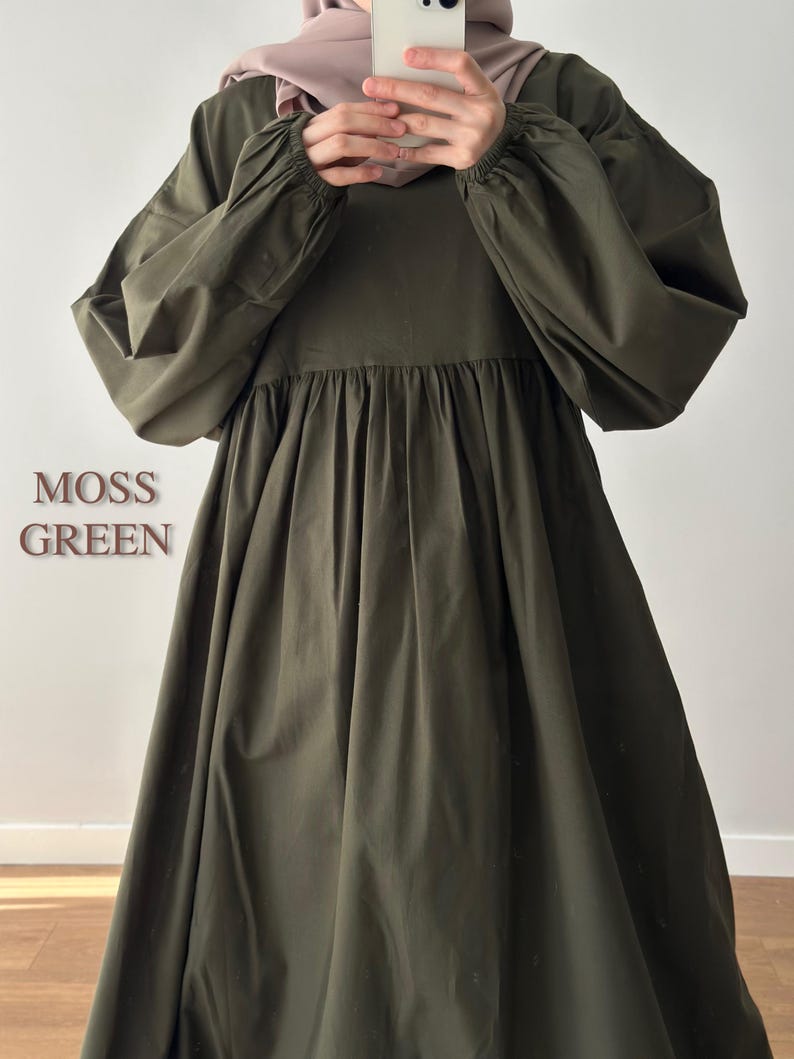 May include: A moss green dress featuring long, voluminous sleeves with elasticated cuffs and a gathered, flowing skirt. The text MOSS GREEN is visible on the left side. The garment has a round neckline and a loose, relaxed fit throughout the bodice.