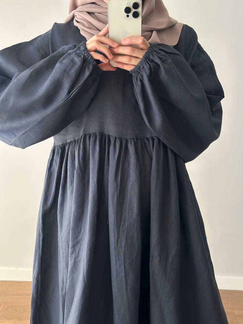 May include: A dark charcoal gray dress featuring long, voluminous sleeves with elasticized cuffs and a gathered, flowing skirt. The garment has a round neckline and a relaxed, loose-fitting silhouette that extends downward from the bodice.