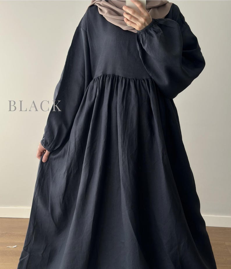 May include: A long-sleeved black dress with a gathered waistline and a full, flowing skirt. The garment features a round neckline and elasticized cuffs on the sleeves. The word BLACK is printed in a light gray serif font on the left side.