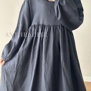 May include: A long-sleeved dress in a dark charcoal gray color, featuring a gathered waistline and elasticized cuffs. The fabric has a smooth, matte finish. The word ANTHRACITE is printed in white capital letters across the chest area of the dress.