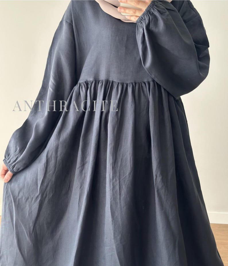 May include: A long-sleeved dress in a dark charcoal gray color, featuring a gathered waistline and elasticized cuffs. The fabric has a smooth, matte finish. The word ANTHRACITE is printed in white capital letters across the chest area of the dress.