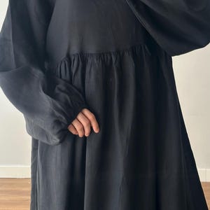 May include: A long-sleeved black dress featuring a gathered waistline and elasticized cuffs. The dress is paired with a solid mauve headscarf that covers the head and neck area, creating a simple and cohesive look with long, flowing fabric.
