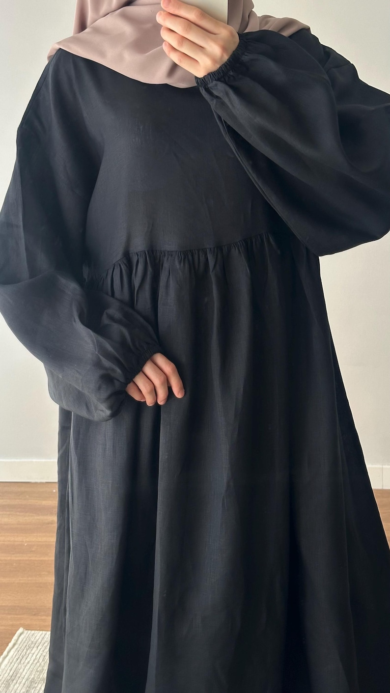 May include: A long-sleeved black dress featuring a gathered waistline and elasticized cuffs. The dress is paired with a solid mauve headscarf that covers the head and neck area, creating a simple and cohesive look with long, flowing fabric.