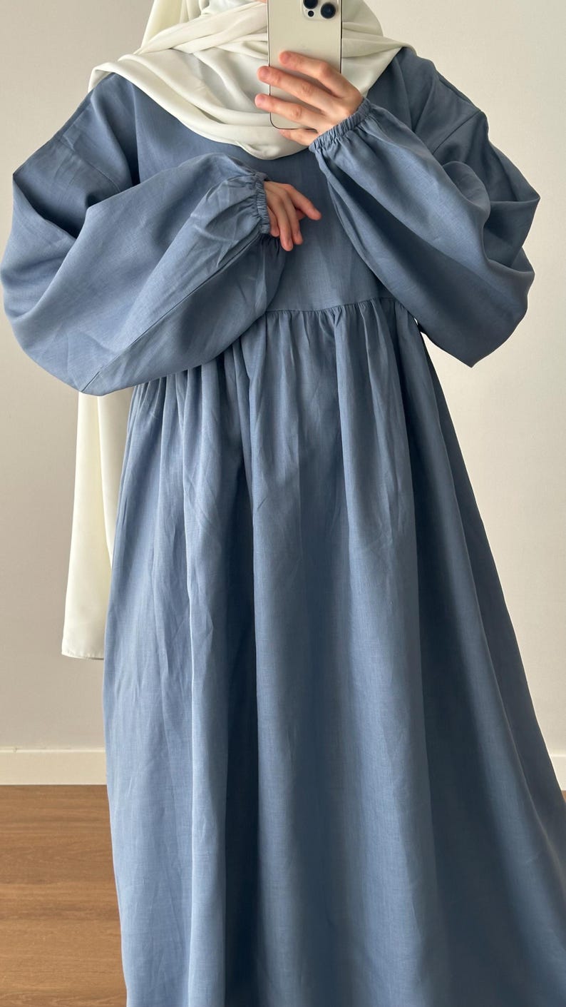May include: A dusty blue long-sleeved dress featuring a gathered waistline and elasticized cuffs, paired with a solid white headscarf. The dress has a loose, flowing silhouette with a round neckline and a soft, matte fabric texture throughout.