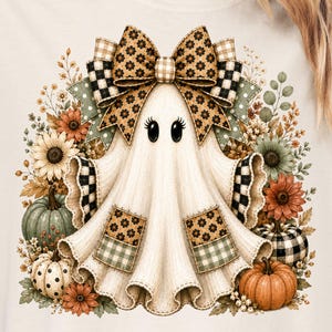 May include: A cream-colored t-shirt featuring a graphic of a ghost with large eyes, wearing a large patterned bow. The ghost is surrounded by sunflowers, autumn leaves, and pumpkins in shades of orange, green, and brown with checkered accents.