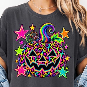 Könnte beinhalten: Ein anthrazitfarbenes T-Shirt mit einer leuchtend bunten Kürbisgrafik im Leopardenmuster. Der Kürbis hat einen regenbogenfarbenen Stiel und ein Blatt, umgeben von hellen, neonfarbenen Sternen in Pink, Gelb, Orange und Blau.