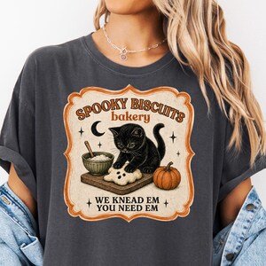 Könnte beinhalten: Ein anthrazitfarbenes T-Shirt mit einer Grafik eines schwarzen Kätzchens, das Teig auf einem Holzbrett neben einer Schüssel und einem Kürbis knetet. Der Text lautet SPOOKY BISCUITS bakery WE KNEAD EM YOU NEED EM, umrahmt von einem orangefarbenen Rand.