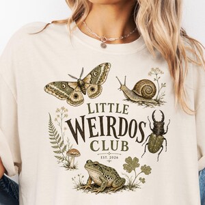 Könnte beinhalten: Ein cremefarbenes T-Shirt mit einem Grafikdruck, der einen Nachtfalter, eine Schnecke, einen Käfer und einen Frosch zeigt, umgeben von botanischen Elementen. Der Text "LITTLE WEIRDOS CLUB EST. 2026" ist mittig in dunkelgrüner Schrift gedruckt.
