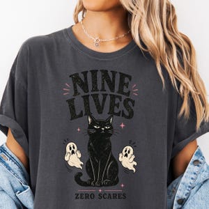 Könnte beinhalten: Ein anthrazitfarbenes T-Shirt mit einem Grafikdruck einer schwarzen Katze, die in der Mitte sitzt und von zwei kleinen weißen Geistern flankiert wird. Über der Katze steht der Text NINE LIVES in Schwarz, darunter der Text ZERO SCARES in Schwarz.