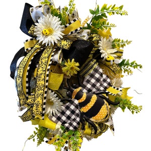 May include: A black and white checkered wreath with yellow and black ribbon, white daisies, and a large black and yellow bee. The wreath is decorated with green leaves and small yellow bees.