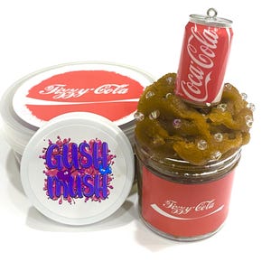 May include: A jar of brown slime topped with a miniature Coca-Cola can and clear beads. The jar has a red label with the text "Fizzy-Cola". Two white containers with the text "Fizzy-Cola" and "Gush Mush" are also in the image.