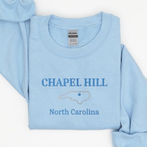 Może przedstawiać: Jasnoszary sweter z napisem "CHAPEL HILL" i zarysowaniem stanu Karolina P&oacute;łnocna z gwiazdą w środku. Napis "North Carolina" znajduje się pod zarysowaniem stanu.