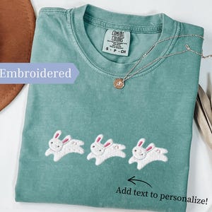 May include: A seafoam green t-shirt with three embroidered white bunnies with pink ears. The shirt has the text "Add text to personalize!" and "Embroidered". A silver necklace with a pendant is on the shirt.
