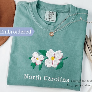 May include: A sage green t-shirt featuring an embroidered design of two white dogwood flowers with yellow centers and green leaves. The text North Carolina is embroidered below the flowers. The shirt has a Comfort Colors label at the neck.