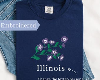 May include: Navy blue t-shirt with an embroidered floral design in purple and green. The word "Illinois" is below the floral design. The shirt is folded on top of a pair of blue jeans. The text "Embroidered" is on the left side of the shirt.