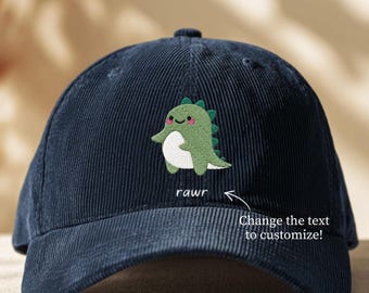 May include: A navy blue corduroy baseball cap featuring an embroidered green dinosaur with a white belly and pink cheeks. Below the dinosaur, the word "rawr" is embroidered in white, with an arrow pointing to it and the text "Change the text to customize!"