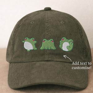 May include: Olive green corduroy baseball cap featuring three embroidered green frog designs with white bellies and red accents. The cap has a curved brim and a top button. Text on the image says "Add text to customize!"