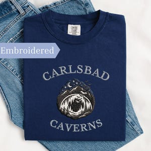Peut inclure: T-shirt bleu marine avec broderie "Carlsbad Caverns". Le motif présente une illustration de grotte avec un ciel nocturne et une lune. Le t-shirt est présenté sur un jean bleu. Le mot "Embroidered" est dans un encadré bleu clair.